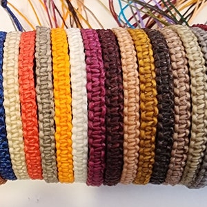 May include: A collection of colourful braided bracelets made with different coloured cords. The bracelets are all about 1.2 cm wide and are tied together in a bundle.