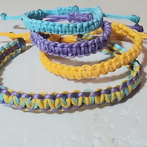 May include: A selection of macrame bracelets in blue, purple, and yellow. Each bracelet has an adjustable sliding knot. The bracelets are approximately 1.3 cm wide. The bracelets are handmade.