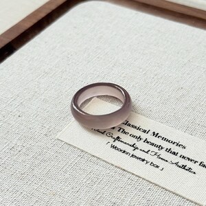 Peut inclure: Une bague violet clair avec un design arrondi. La bague est faite d'un matériau translucide et repose sur une étiquette blanche avec du texte noir. L'étiquette indique "Classical Memories". La bague est sur une surface texturée blanc cassé.