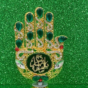 May include: A gold-colored decorative hand with green and clear gemstones. The hand-shaped ornament has a green velvet center with Arabic script. The base is gold-colored and the background is a glittery green.