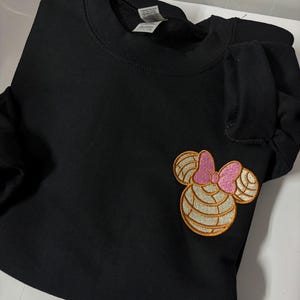 May include: Black sweatshirt with a brown and pink embroidered Minnie Mouse design on the left side of the chest.