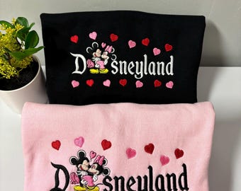 May include: Two pink and black sweatshirts with an embroidered image of Mickey Mouse and Minnie Mouse kissing. The text "Disneyland" is embroidered below the image. The sweatshirts are decorated with pink and red hearts.