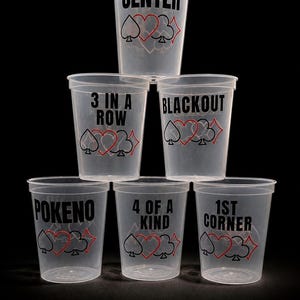 May include: A stack of clear plastic cups with black text and red suit symbols. Each cup has a different phrase: "CENTER", "3 IN A ROW", "BLACKOUT", "POKENO", "4 OF A KIND", and "1ST CORNER". The cups are arranged against a black background.