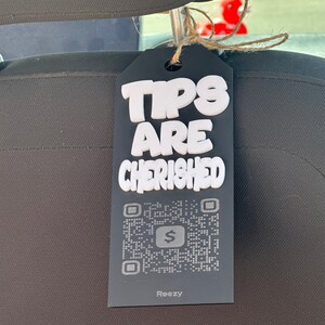 May include: A black tag with the words "TIPS ARE CHERISHED" in white, 3D-style lettering. Below the text is a QR code and a dollar sign icon. The tag is attached to a car seat headrest with twine.