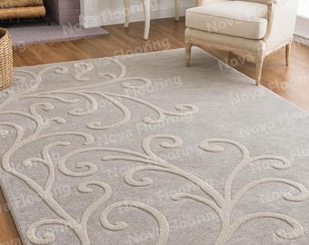May include: A rectangular, light gray area rug with a beige, stylized floral pattern. The rug is placed on a hardwood floor, and a white chair and side table are visible in the background. The rug is a decorative element for a living room.