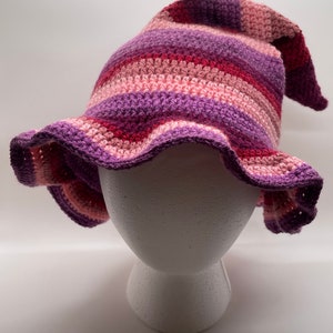 May include: A handmade crochet hat in shades of purple, pink, and burgundy. The hat has a pointed top and a wide, ruffled brim. The yarn is striped, creating a textured effect. The hat is displayed on a white mannequin head.