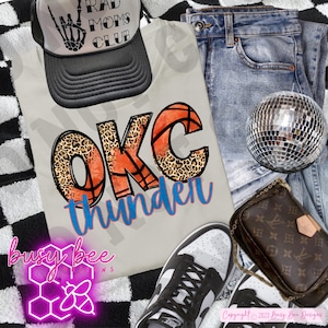 May include: A cream-colored t-shirt with "OKC Thunder" text and basketball graphics, paired with a black trucker hat that says "Rad Moms Club." The image includes blue jeans, a disco ball, a small brown purse, and black and white sneakers.