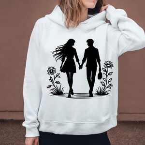 Może przedstawiać: Biała bluza z kapturem z czarną sylwetką pary trzymającej się za ręce i idącej przez pole kwiat&oacute;w.