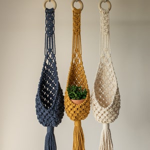 May include: Three macrame plant hangers with wooden rings, each in a different colour: navy blue, mustard yellow, and natural white. The yellow hanger has a small potted plant inside.