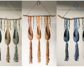 May include: Three macrame plant hangers with a wooden branch at the top. The hangers are in different colors: green, beige, and blue.