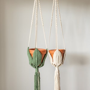 May include: Two macrame plant hangers with terracotta pots. One hanger is green and the other is natural white.