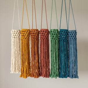 May include: Seven macrame plant hangers in a variety of colours, including white, yellow, orange, red, green, blue, and light blue. Each hanger features a knotted design and a fringe at the bottom.