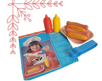 Puede incluir: Un juego de comida de juguete Fisher-Price con una botella exprimible roja y amarilla, una espátula de plástico azul y una bandeja rosa con cuatro perritos calientes. El juego incluye instrucciones para hacer perritos calientes.