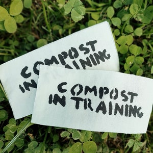 May include: Two white rectangular labels with the text "COMPOST IN TRAINING" in black stencil font are displayed on a bed of green clover and grass. The labels are slightly overlapping.