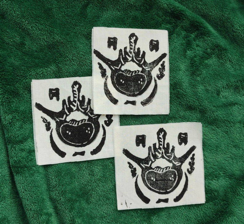 Puede incluir: Tres parches cuadrados de tela blanquecina con una impresi&oacute;n en lin&oacute;leo negro de una imagen anat&oacute;mica estilizada. El dise&ntilde;o presenta una forma central redondeada con l&iacute;neas radiales y elementos rectangulares m&aacute;s peque&ntilde;os. Los parches est&aacute;n dispuestos sobre una superficie verde texturizada.