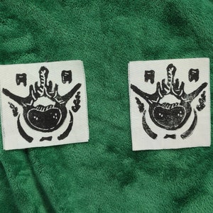 Puede incluir: Dos parches de tela blanca con un dise&ntilde;o anat&oacute;mico negro que representa una pelvis y una columna vertebral, con elementos decorativos. Los parches se presentan sobre un fondo verde.