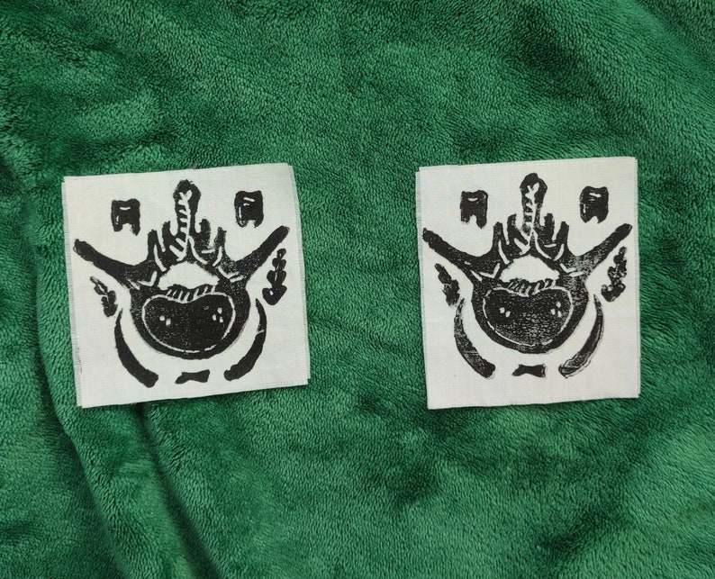 Puede incluir: Dos parches de tela blanca con un dise&ntilde;o anat&oacute;mico negro que representa una pelvis y una columna vertebral, con elementos decorativos. Los parches se presentan sobre un fondo verde.