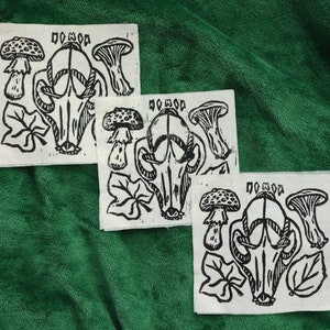 Puede incluir: Tres parches de tela blanca con un diseño de linograbado negro. Cada parche presenta una calavera, setas y hojas, con la palabra "DOMOD" impresa en la parte superior. Los parches están dispuestos sobre un fondo verde texturizado.