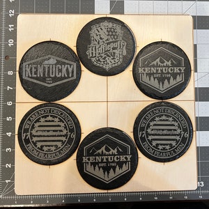 May include: A collection of six black slate coasters with engraved designs. The coasters feature text and graphics, including the word "Kentucky" with the year "1792", the Hufflepuff crest, and the phrase "We are not descended from fearful men."