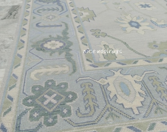 May include: A light blue and ivory-colored rug with a detailed floral and geometric pattern. The rug features a border with repeating shapes and the text "rice wool rugs". The rug has a fringe along the bottom edge.