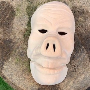 Puede incluir: Una máscara de cerámica blanca con un hocico de cerdo y rasgos humanos. La máscara tiene una superficie texturizada y está sentada sobre una superficie de madera.