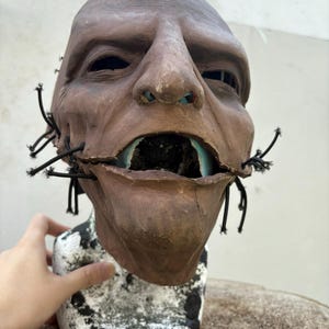 May include: A brown, grotesque mask with an open mouth revealing blue and black details. The mask has a weathered appearance, with black string-like elements attached around the mouth. The mask is mounted on a white and black pedestal.