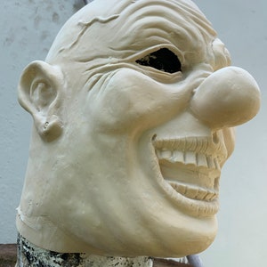 May include: A close-up of a white, rubber clown mask with a large, exaggerated smile and a red, bulbous nose. The mask is made of a soft, flexible material and has a realistic texture.