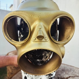 Puede incluir: Una máscara de gas de color dorado con dos grandes piezas redondas para los ojos y una pieza redonda más pequeña para la nariz. La máscara tiene una superficie texturizada y parece estar hecha de plástico.