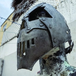 May include: A dark gray, medieval-style helmet with riveted plates and a visor featuring rectangular eye slits. The helmet is displayed on a mannequin head, showcasing its textured surface and leather strap.