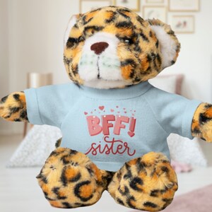 May include: A plush leopard-print teddy bear wearing a light blue shirt with the text "BFF! sister" in pink. The bear has a brown nose and white accents on its ears.
