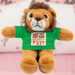 Könnte beinhalten: Ein Stofftier-Löwe mit brauner Mähne und beigefarbenem Fell, das ein grünes T-Shirt trägt. Auf dem Shirt steht der Text "DOES THIS SHIRT MAKE ME LOOK FAT?" im Retro-Stil. Der Löwe hat einen freundlichen Gesichtsausdruck.
