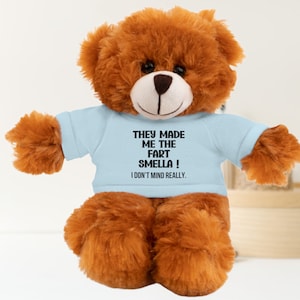 May include: A brown teddy bear wearing a light blue t-shirt with the text "They made me the fart smella! I don't mind really."