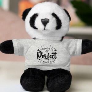 Puede incluir: Un peluche de panda con pelaje blanco y negro, vistiendo una camiseta gris. La camiseta presenta el texto "Nobody is Perfect, I am Nobody" en una fuente negra y decorativa. El panda tiene patas y orejas negras.