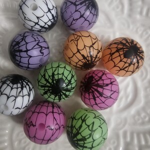 Peut inclure: Un assortiment de perles rondes de différentes couleurs : blanc, violet, orange, vert et rose. Chaque perle est ornée d'un motif de toile d'araignée noire et d'une araignée noire, parfait pour les créations d'Halloween.