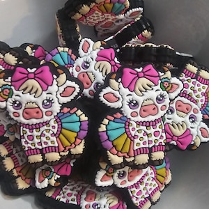 May include: A group of colorful cow charms with pink bows, white polka dots, and rainbow-colored skirts. The charms are made of rubber and are designed to be used as shoe charms or keychains.
