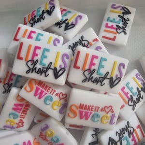 May include: A collection of colourful silicone charms with the text "Life is Short" and "Make it Sweet" in various colours and fonts.