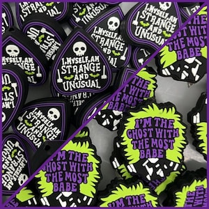 May include: A collection of black and green charm-like pendants with various spooky sayings. Some of the charms feature a skull and crossbones, while others feature a ghost with green hair. The text on the charms includes "I myself am strange and unusual" and "I'm the ghost with the most babe".