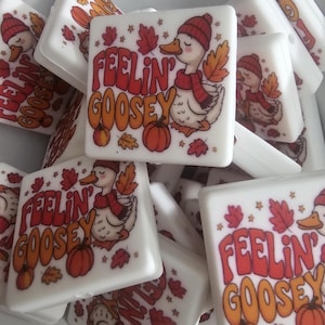 May include: White square tiles with a cartoon duck wearing a red hat and scarf, surrounded by fall leaves and pumpkins. The text "FEELIN' GOOSEY" is in orange and red lettering. The tiles are likely decorative or for crafts.