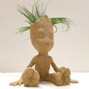 Pode incluir: Um vaso de cerâmica marrom em forma do personagem Groot da série de filmes Guardiões da Galáxia. O vaso tem uma pequena planta crescendo no topo de sua cabeça.