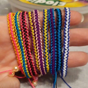Könnte beinhalten: Eine Sammlung bunter Freundschaftsarmbänder. Jedes Armband hat ein gewebtes Design in verschiedenen Farben, darunter Rosa, Gelb, Blau und Lila. Einige Armbänder haben Perlenakzente. Die Armbänder werden zusammen präsentiert.