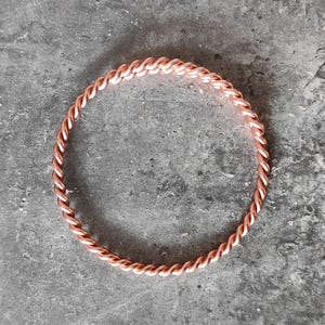 Op de afbeelding: Een koperkleurige armband met een gedraaid touwontwerp. De armband is cirkelvormig en ligt op een grijs, getextureerd oppervlak. Het metaal heeft een warme, roodachtig bruine tint.