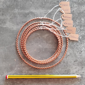 May include: A set of five copper wire hoops, each with a different tag attached. The tags are brown paper with white string and have the words "NOW", "LIGHT", "HARMONY", "SACRED", and "POWER" written on them. A yellow pencil is lying below the hoops.