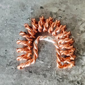 May include: A handcrafted copper-colored wire hair accessory, shaped like a half-circle. The piece is made of twisted wire, with a silver-colored metal band at the base. The accessory is displayed on a gray surface.