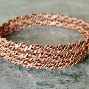 Puede incluir: Una pulsera de color cobre con un diseño de cuerda trenzada. La pulsera está hecha de tres hebras de metal trenzado, creando un patrón texturizado e intrincado. La joya se exhibe sobre una superficie gris.