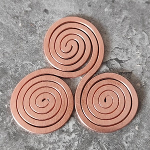 May include: Three copper spiral shapes arranged in a trefoil pattern. Each spiral is a flat, circular design with a central point and concentric rings. The copper has a warm, reddish-brown hue, and the background is a textured gray surface.