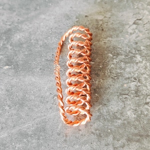 May include: A copper-colored, coiled wire, possibly for jewelry making or crafts, lies on a gray surface. The wire is tightly wound in a spiral pattern, with a braided section extending from the coil. The overall appearance is metallic and textured.