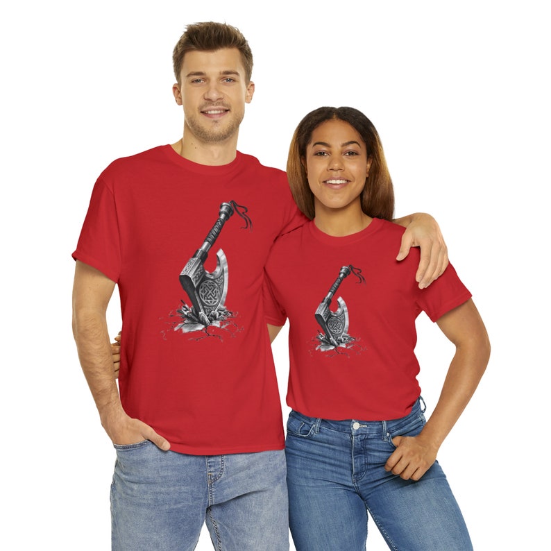 May include: A bright red short-sleeved t-shirt featuring a graphic print of a silver battle axe embedded in a cracked stone surface. The axe head displays intricate Celtic knotwork patterns, and a small black ribbon is tied to the handle.