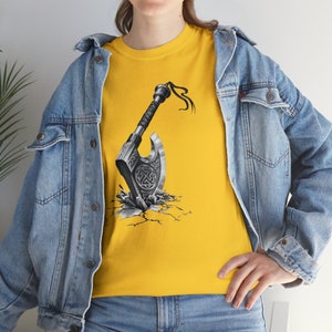 May include: A bright yellow t-shirt featuring a graphic print of a silver battle axe with intricate Celtic knotwork patterns on the blade. The axe appears to be embedded in a cracked surface, and the shirt is worn under a light blue denim jacket.