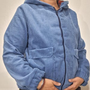 Könnte beinhalten: Eine blaue Cordjacke mit Reißverschluss und Kapuze. Die Jacke hat eine Tasche auf der Vorderseite und einen gerippten Bund.