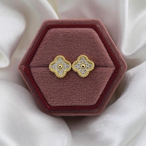 May include: A pair of gold-colored clover-shaped stud earrings with a central gold bead and surrounding clear stones. The earrings are displayed in a maroon velvet jewelry box, set against a backdrop of white fabric.